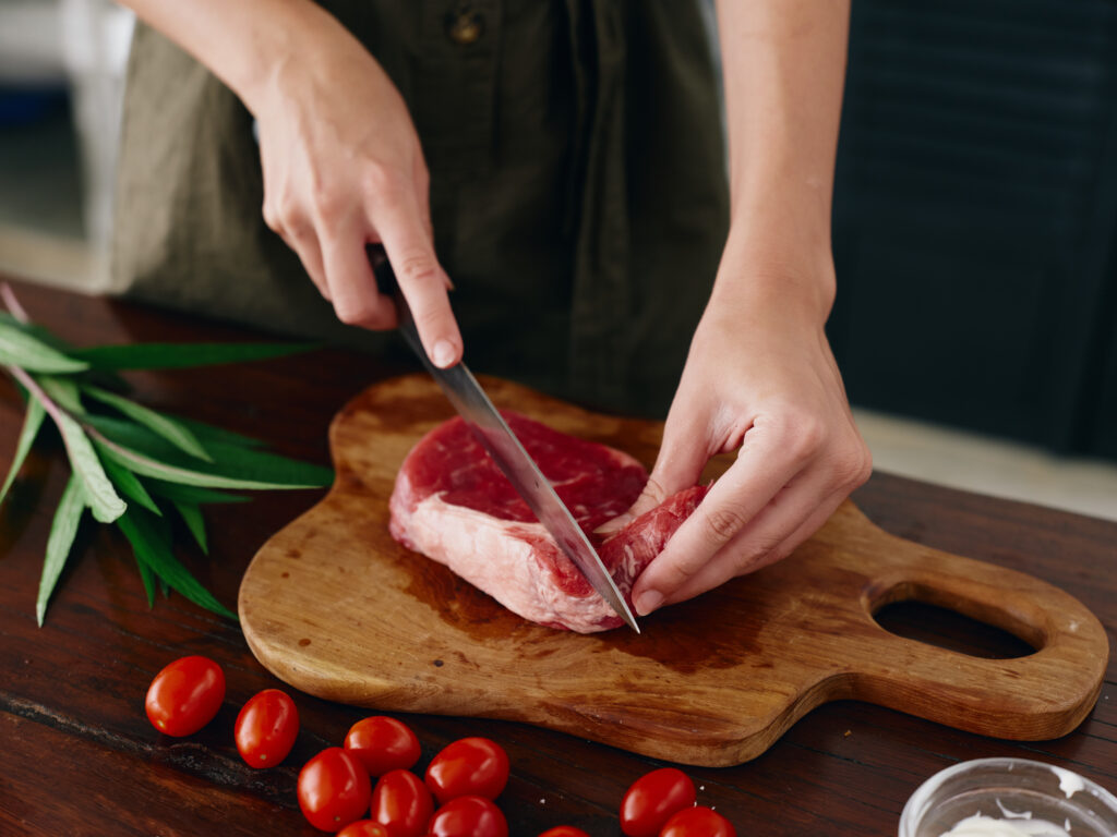 Woman with knife in hand cutting fresh steak meat for roasting in kitchen with salt pepper and other spices on table, red cherry tomatoes and herbs, preparing dinner. High quality photo